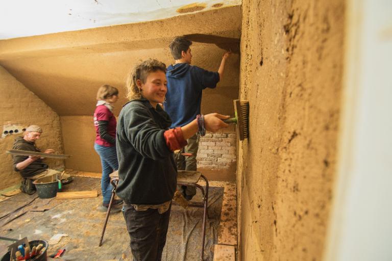 Workcamp-Teilnehmende gestalten eine Innenraumwand mit Lehm. | Foto: Hendrik Silbermann (ARTWORKs)